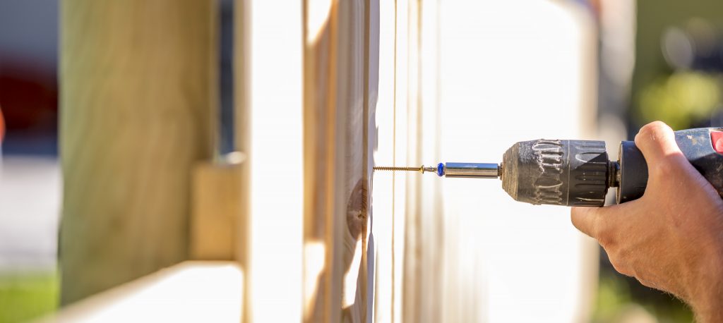 Man erecting a wooden fence outdoors using a handheld electric drill to drill a hole to attach an upright plank, close up of his hand and the tool in a DIY concept.